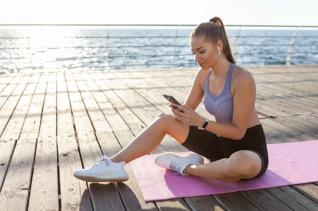 Slm fit mujer en ropa deportiva usa un teléfono inteligente mientras está sentado en una colchoneta en la playa al amanecer.