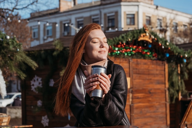 Slow living en el entorno de la ciudad, Urban Slow life, mindfulness, disfrutar del momento, conciliación de la vida laboral y personal. Mujer joven pelirroja disfrutando del sol y bebiendo café para llevar en las calles de la ciudad de invierno.