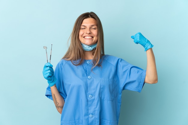 Slowakischer Zahnarzt, der Werkzeuge hält, die auf blaue Wand lokalisiert werden, die starke Geste tut