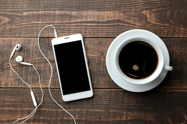 Smartphone com fones de ouvido e uma xícara com café em uma mesa de madeira marrom. vista de cima