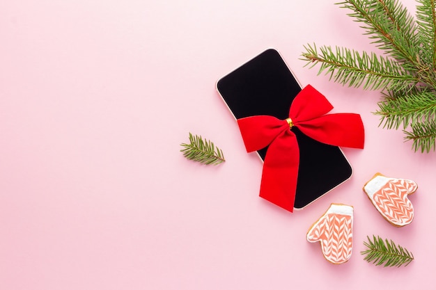 Smartphone com laço vermelho para um presente de Natal em fundo rosa perto de abeto e biscoitos