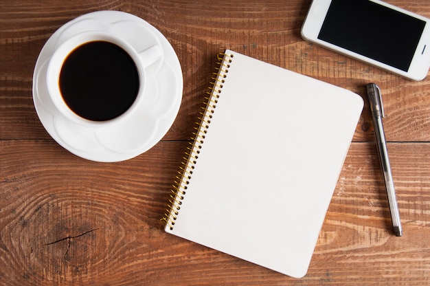 Smartphone com notebook e xícara de café sobre fundo de madeira.