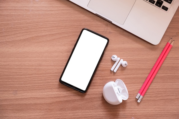 Smartphone com tela em branco e fones de ouvido sem fio e laptop na mesa de madeira