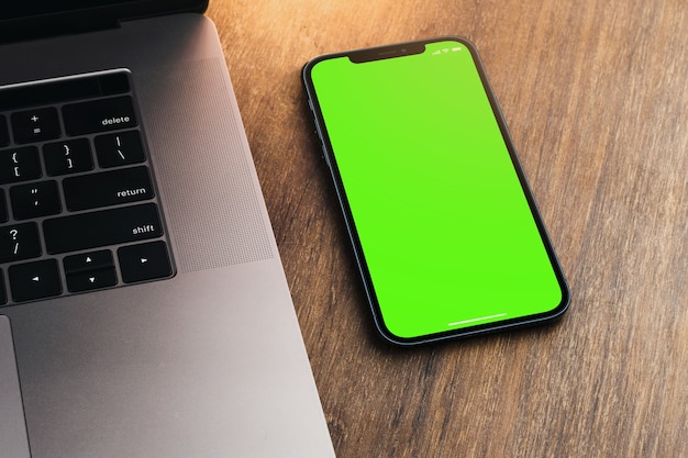 Smartphone de tela verde em branco sobre fundo de madeira com um computador ao lado. Chave de croma.