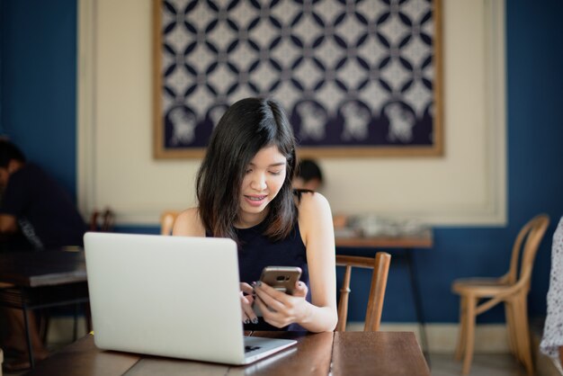 Smartphone de utilização autônomo da menina asiática ao trabalhar com seu portátil no coffeeshop.