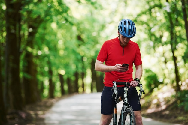 Smartphone halten. Radfahrer auf einem Fahrrad ist auf der Asphaltstraße im Wald am sonnigen Tag