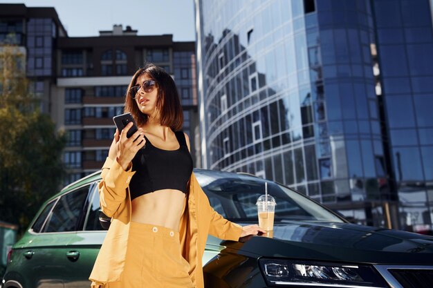 Con smartphone Joven mujer de moda con abrigo de color burdeos durante el día con su coche