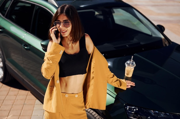 Con smartphone Joven mujer de moda con abrigo de color burdeos durante el día con su coche