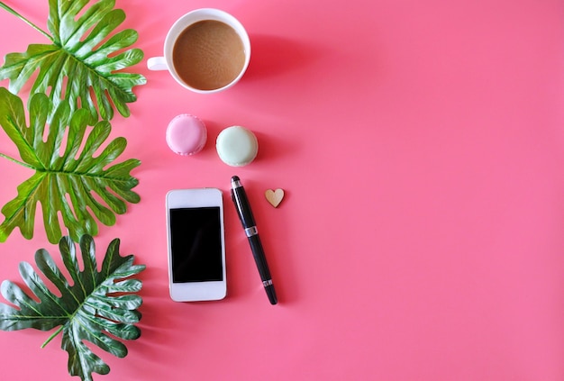 Smartphone, Kaffeetasse, Stift, Macaron und tropisches Blatt auf rosa Hintergrund