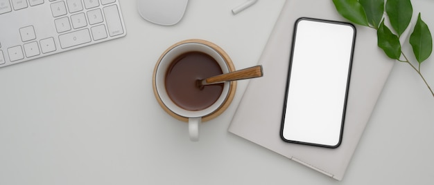 Smartphone mit leerem bildschirm über dem zeitplanbuch auf weißem schreibtisch mit computergeräten und heißer kakaotasse