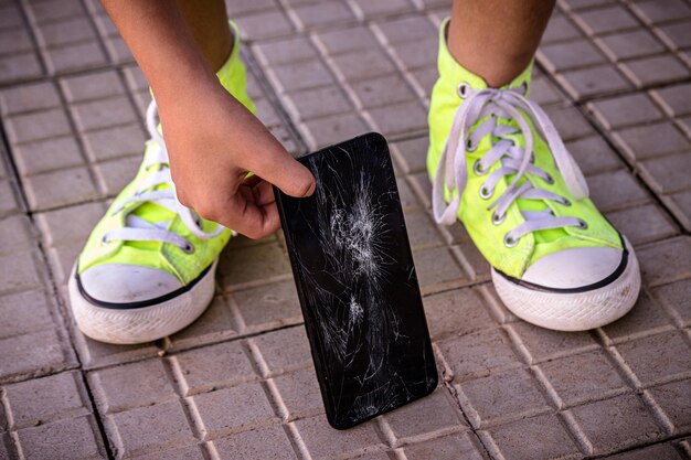 Smartphone mit zerbrochenem Bildschirm, das von einem unbeholfenen jungen Mann vom Boden aufgehoben wurde. Handyfilm im Herbst zerstört