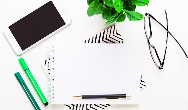 Smartphone, óculos, planta verde, marcadores verdes, caderno em branco e caneta na área de trabalho. Postura plana.