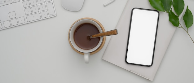 Smartphone de pantalla en blanco sobre el libro de horarios en el escritorio de oficina blanco con dispositivos informáticos y taza de chocolate caliente