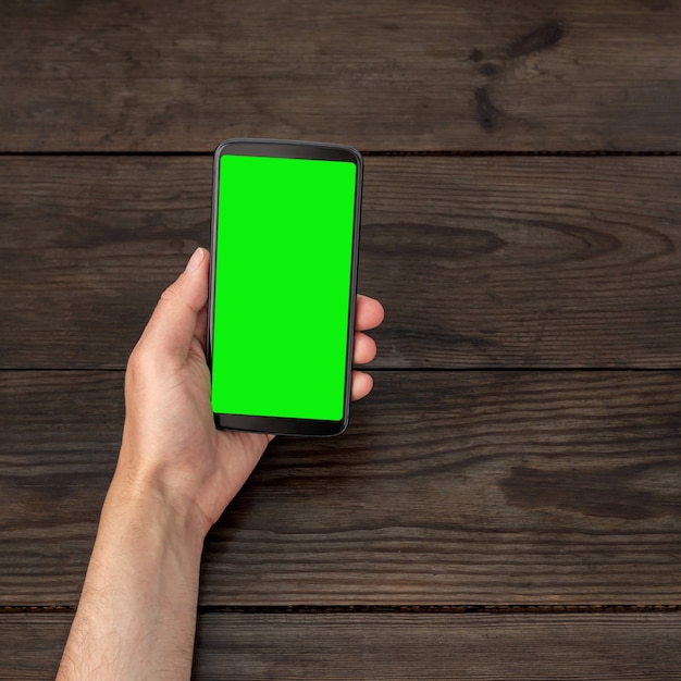Smartphone con una pantalla verde en la mano sobre un fondo de mesa de madera.