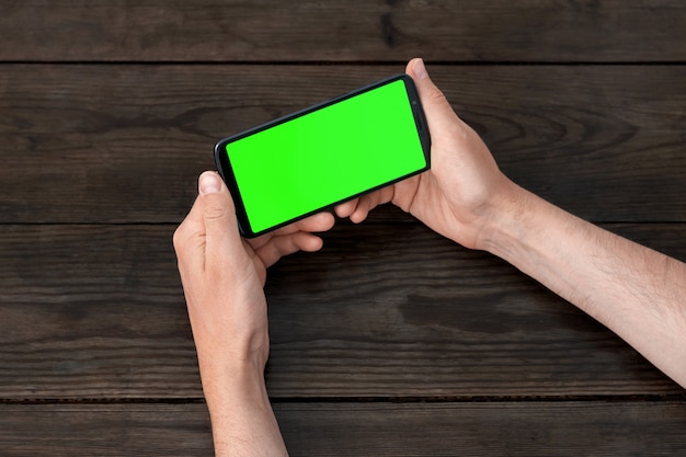 Smartphone con pantalla verde en manos, posición horizontal, sobre una mesa de madera.