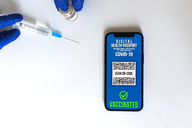 Smartphone con pasaporte de vacuna en la mesa del médico, mano en guantes con jeringa y frasco de vacuna COVID-19, fondo blanco, concepto de viaje seguro