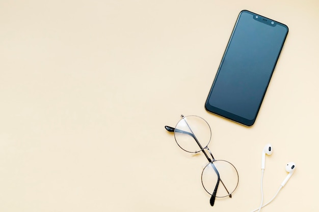 Smartphone preto com tela em branco e fones de ouvido com fio, óculos celular em fundo bege