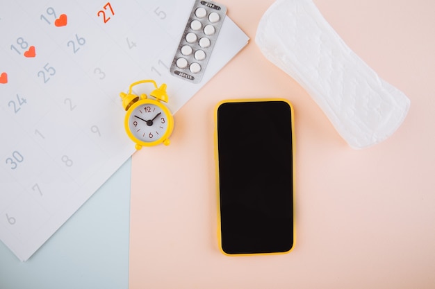 Smartphone para rastrear su ciclo menstrual y para marcas. PMS y el concepto de días críticos. Tampón de algodón, compresa diaria y alarma amarilla sobre fondo rosa.