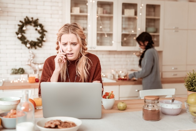 Smartphone tragen. Verwirrte und unglückliche blonde Frau, die ernsthaften Telefonanruf beim Beobachten von Informationen auf Laptop hat