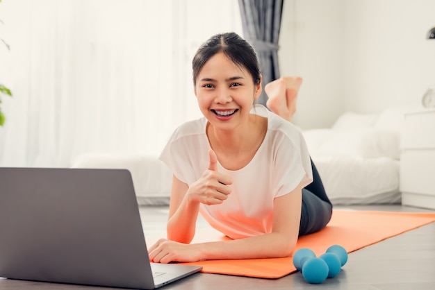 Smiley junge asiatische Frau auf einer Yogamatte und hebt ihre Hand, um wie Pose auf dem Schlafzimmer zu zeigen.