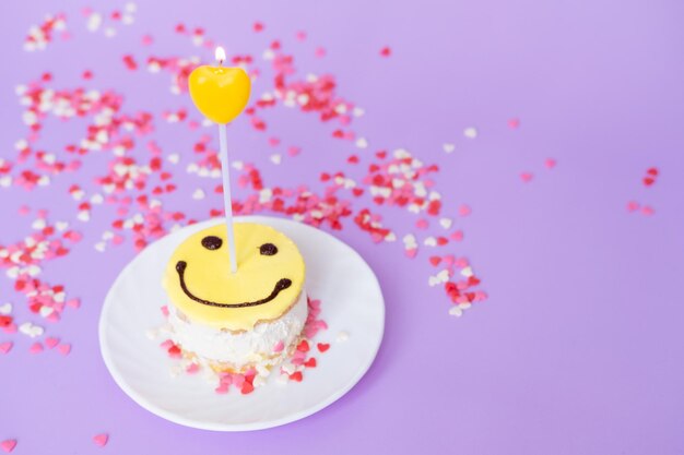 Smiley de pastel amarillo con una vela en forma de corazón sobre un fondo morado Concepto de día de San Valentín y cumpleaños