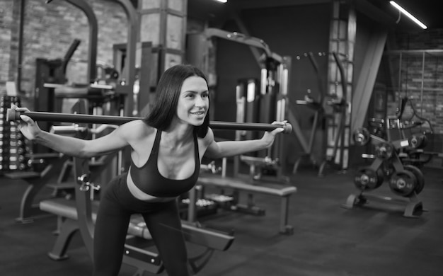 Smilling joven morena haciendo ejercicio gimnástico con barra para el cuerpo sobre sus hombros inclinando su cuerpo en el gimnasio