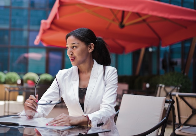 Smilling mujer de negocios afro lista para la entrevista mientras está sentado a la mesa en la cafetería al aire libre