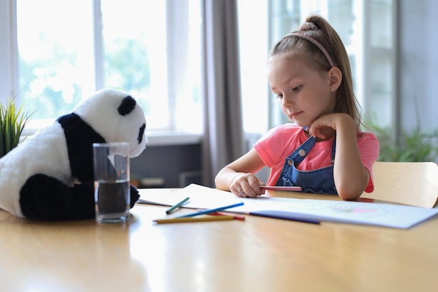 Smilling niña feliz sentada en la mesa con un oso panda de juguete cerca de ella disfrutando de la actividad creativa, dibujando lápices para colorear en álbumes.