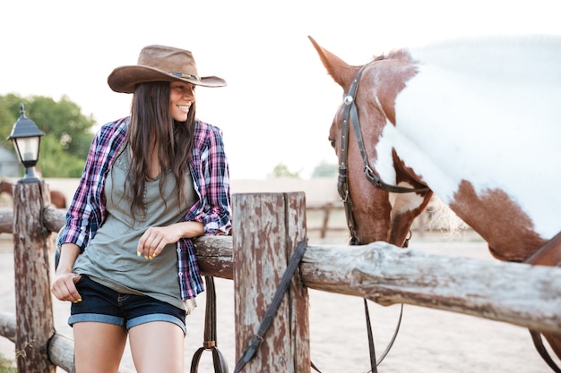 Smilng bastante joven vaquera de pie con su caballo en el rancho