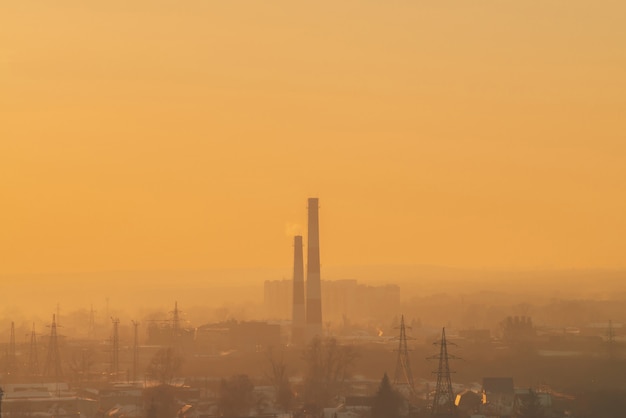 Smog unter Silhouetten von Gebäuden bei Sonnenaufgang. Schornstein im Morgenhimmel. Umweltverschmutzung bei Sonnenuntergang. Schädliche Dämpfe vom Stapel über der Stadt.