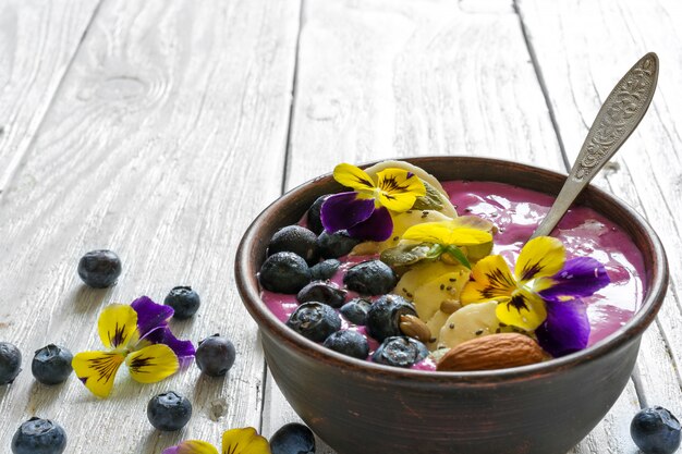 Smoothie-schüssel mit einem löffel mit frischen beeren, bananen, chiasamen, nüssen und blumen für ein gesundes frühstück