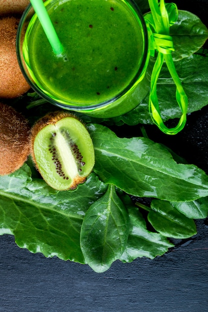 Smoothie verde perto de ingredientes para ele, sobre fundo preto de madeira. Kiwi e espinafre.