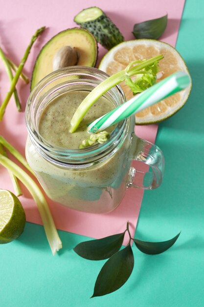 Foto smoothies saudáveis vegetarianos de vegetais verdes com folhas verdes, fatias de limão, abacate, coco e canudo plástico em uma tigela de vidro com fundo de papel verde rosa duotônico.