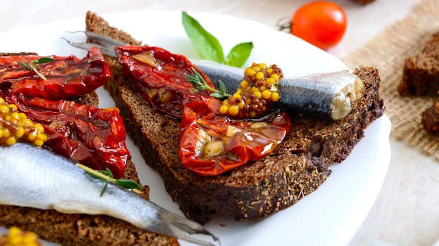 Smorrebrod sabroso en un plato blanco. Sándwiches con pan de centeno negro, tomates secos, anchoas saladas, mostaza.