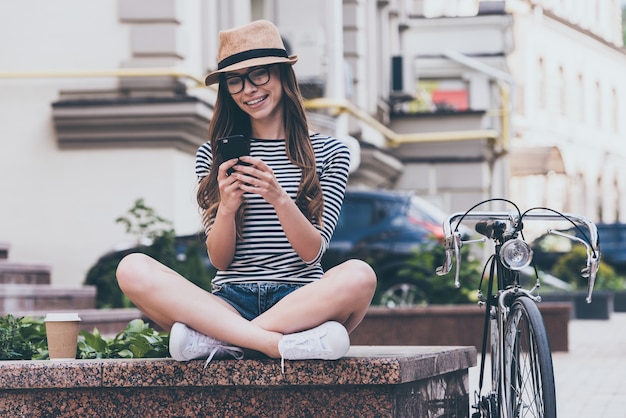 SMS an Freund. Schöne junge lächelnde Frau, die ihr Smartphone anschaut, während sie auf der Bank in der Nähe ihres Fahrrads sitzt
