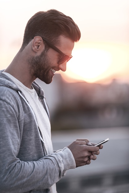 SMS an Freund. Seitenansicht eines gutaussehenden jungen Mannes mit Sonnenbrille, der Handy hält und lächelt, während er im Freien steht