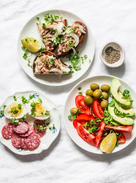 Snack Vorspeisen Tapas Frühstückstisch Salami Makrele Frischkäse Sandwiches Oliven Avocado Tomaten Mikrogemüse auf hellem Hintergrund Draufsicht