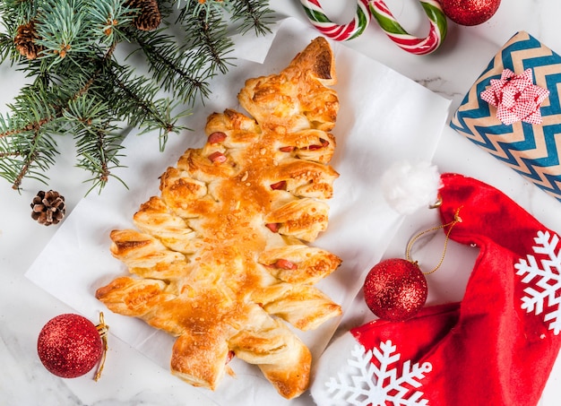 Snacks für die Weihnachtsfeier. Geschlossene Pizza aus Blätterteig in Form eines Weihnachtsbaumes. Auf einem weißen Marmortisch. Mit Weihnachtsschmuck. Ansicht von oben