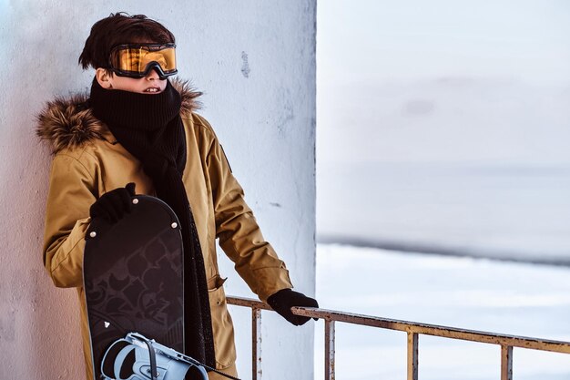 Un snowboarder adolescente vestido con ropa abrigada y gafas protectoras sentado en una baranda cerca de la playa nevada