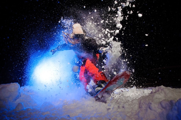 Snowboarder ativa vestida com uma roupa esportiva laranja, pulando na encosta da montanha à noite sob a luz azul