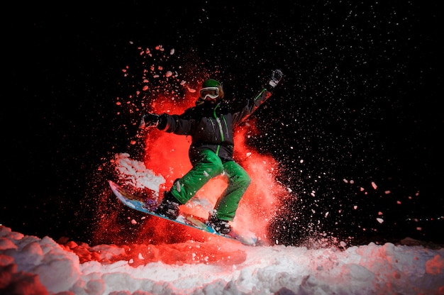 Snowboarder ativa vestida com uma roupa esportiva verde, pulando sob a neve durante a noite sob a luz vermelha