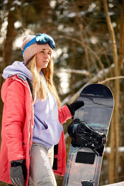 Una snowboarder caucásica sostiene snowboard en un hermoso fondo de bosque nevado en un día soleado