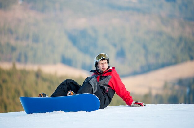 Snowboarder hombre con snowboard sentado en ladera nevada