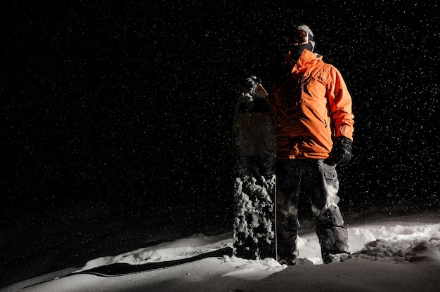 Snowboarder in orange Sportbekleidung und Maske, die mit einem Brett auf schneebedecktem Hügel nachts stehen
