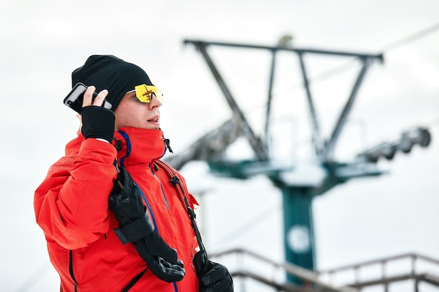 Snowboarder masculino descansando na encosta da colina, falando ao telefone em um lindo dia ensolarado de inverno na estância de esqui. Céu azul, floresta e pistas de esqui com teleférico ao fundo.
