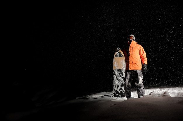 Snowboarder masculino no sportswear laranja em pé com a prancha na neve à noite