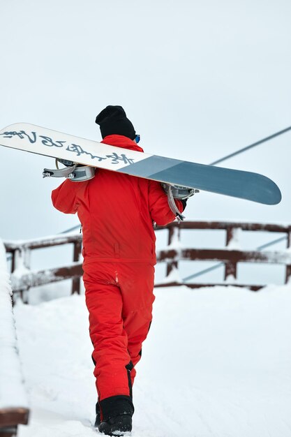 Snowboarder masculino con traje rojo caminando por la colina nevada con concepto de snowboard, esquí y snowboard.