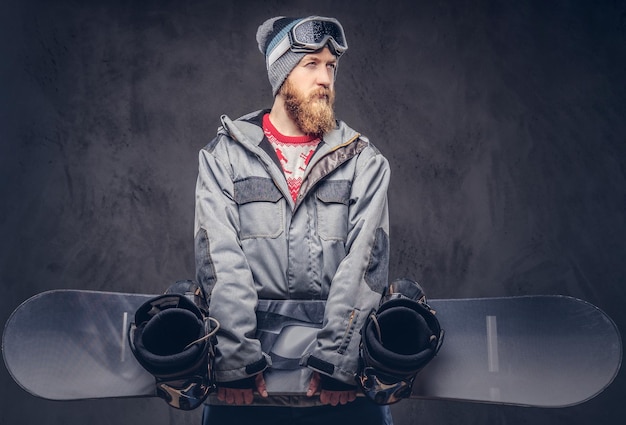 Snowboarder pelirrojo brutal con barba completa en un sombrero de invierno y gafas protectoras vestidas con un abrigo de snowboard sostiene una tabla de snowboard en un estudio. Aislado en un fondo de textura oscura.