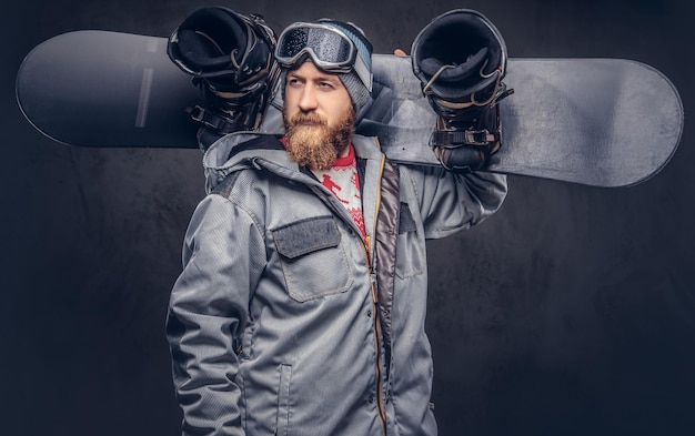 Snowboarder pelirrojo brutal con barba completa en un sombrero de invierno y gafas protectoras vestidas con un abrigo de snowboard sostiene una tabla de snowboard en su hombro en un estudio. Aislado sobre fondo gris.