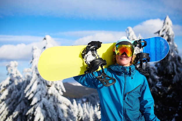 Snowboarder sorridente posando carregando snowboard no ombro na estância de esqui perto da floresta antes do freeride sertão e usando óculos reflexivos roupa de moda colorida Equipamento de snowboard moderno